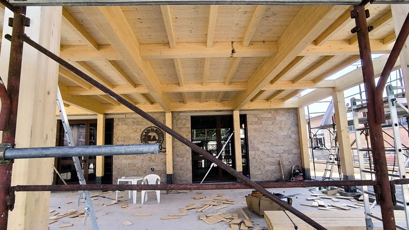 Veranda con struttura di travi in legno lamellare a vista, parete rivestita in pietra e ingresso vetrato, con ponteggi in primo piano durante il cantiere.